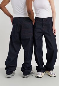Pantalones de mezclilla azul oscuro con un ajuste holgado, que presentan costuras rojas, múltiples bolsillos y un diseño casual. Combinados con zapatillas deportivas.