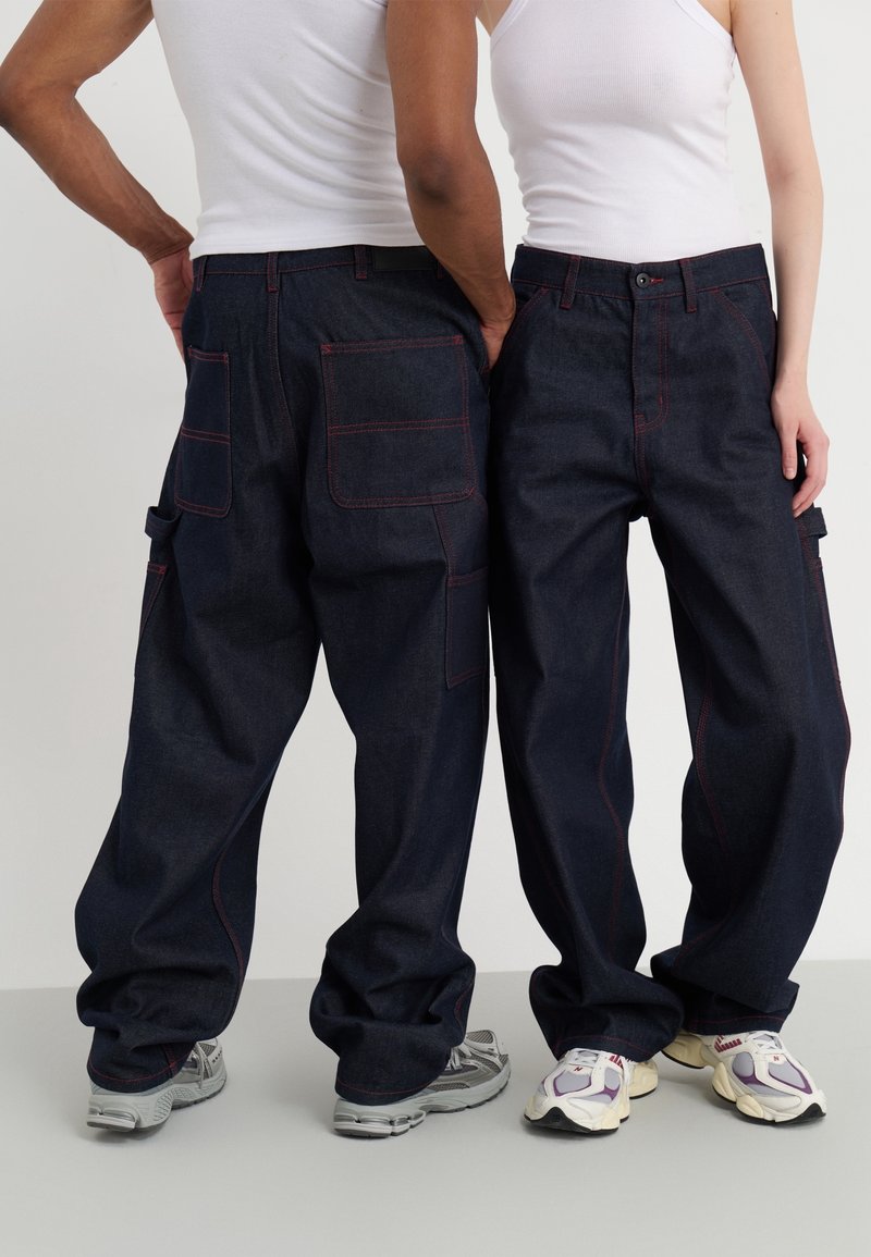 Pantalones de mezclilla azul oscuro con un ajuste holgado, que presentan costuras rojas, múltiples bolsillos y un diseño casual. Combinados con zapatillas deportivas.