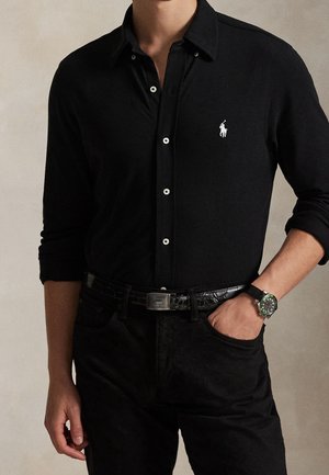 Un mannequin porte une chemise noire boutonnée avec un petit logo blanc sur la poitrine, un pantalon noir, une ceinture et une montre-bracelet.