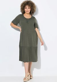 Vestido verde oliva con mangas cortas, un cuerpo fruncido, detalle con cordón y una falda de volantes. Combinado con sandalias de plataforma. Textura de tela suave.