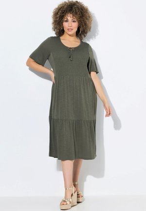 Olivengrünes Kleid mit kurzen Ärmeln, smocktem Oberteil, Zugbanddetails und schichtbarem Rock. Getragen mit Plateausandalen. Glatte Stofftextur.