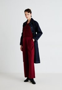 Denim trench coat in dark blue with light stitching, paired with a burgundy knitted top and loose, high-waisted trousers. Black ankle boots.