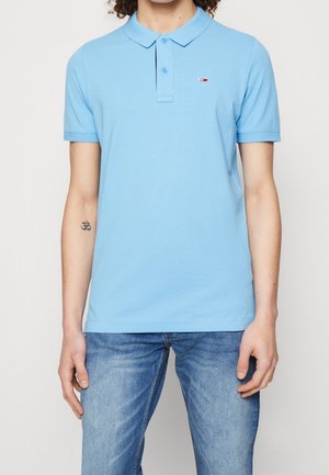 Personne portant un polo bleu clair avec un petit logo sur la poitrine et un jean bleu, se tenant devant un fond blanc uni.