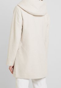 Beige ullkappa med en stor, vikbar huva, långa ärmar och sidofickor. Texturerad tyg med ett subtilt fiskbensmönster.