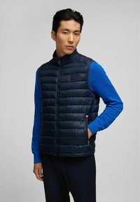 Marineblaues gepolstertes Vest mit hohem Kragen, Reißverschluss und Fronttaschen, kombiniert mit einem leuchtend blauen Langarmshirt. Glatter, gesteppten Textur.