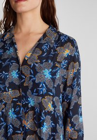 Blusa nera con vivace motivo floreale in blu, arancione e bianco. Presenta un colletto, bottoni e un design con vita arricciata.