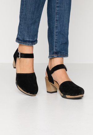 Sabots à talons en velours noir avec une bride de cheville, un talon en bois et un bout rond. La semelle présente des coutures visibles et une finition texturée.