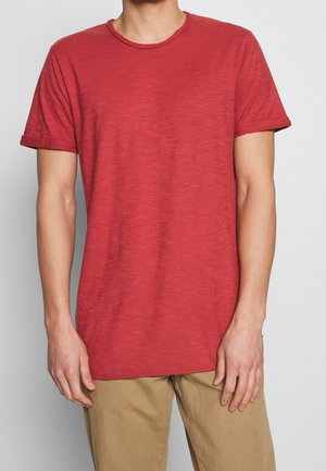 Uomo che indossa una maglietta rossa a maniche corte con scollo rotondo e pantaloni beige, in piedi davanti a uno sfondo grigio chiaro uniforme.