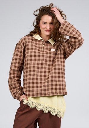 Jeune femme aux cheveux bouclés portant une chemise à carreaux marron sur un haut jaune orné de dentelle, posant avec une main dans les cheveux et un léger sourire.