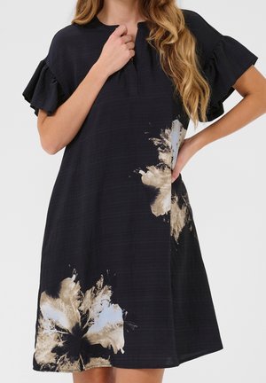 Femme portant une robe noire à manches courtes avec des manches à volants et de grandes impressions florales beige et blanches près de l'ourlet et sur le côté.