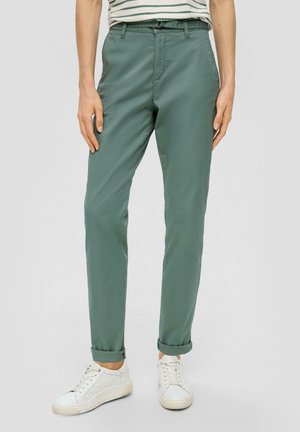 Person iført grønne, smalle bukser med opsmøg, hvide sneakers og en stribet skjorte, stående foran en ensfarvet baggrund.