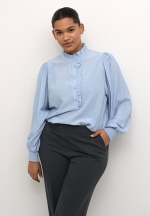 Heled sinine pluus, millel on rätsepakaelus ja nööpidega eest, balloonihargid ja kokku volditud varrukad, koos tumedas hallis istuvate pükstega.