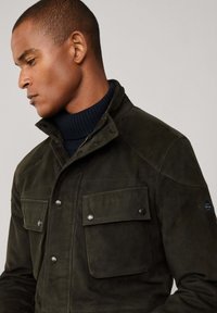 Giacca scamosciata verde scuro con colletto alto, chiusure con bottoni a pressione, due taschini sul petto e una discreta applicazione del logo sulla manica.