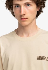 Beige shirt met korte mouwen van katoen, met een bruin logo aan de linkerkant. De stof lijkt zacht en glad met een ronde hals.