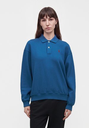 Femme aux cheveux bruns raides portant un polo bleu à manches longues avec un logo rouge et un pantalon noir, debout sur un fond blanc uni.