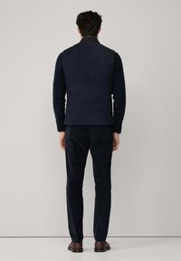 Mornarica prešana prsluk preko crne majice s dugim rukavima, u kombinaciji s tamnim hlačama i smešnim cipelama. Glatka tekstura, minimalistički dizajn, bez vidljivih logoa.