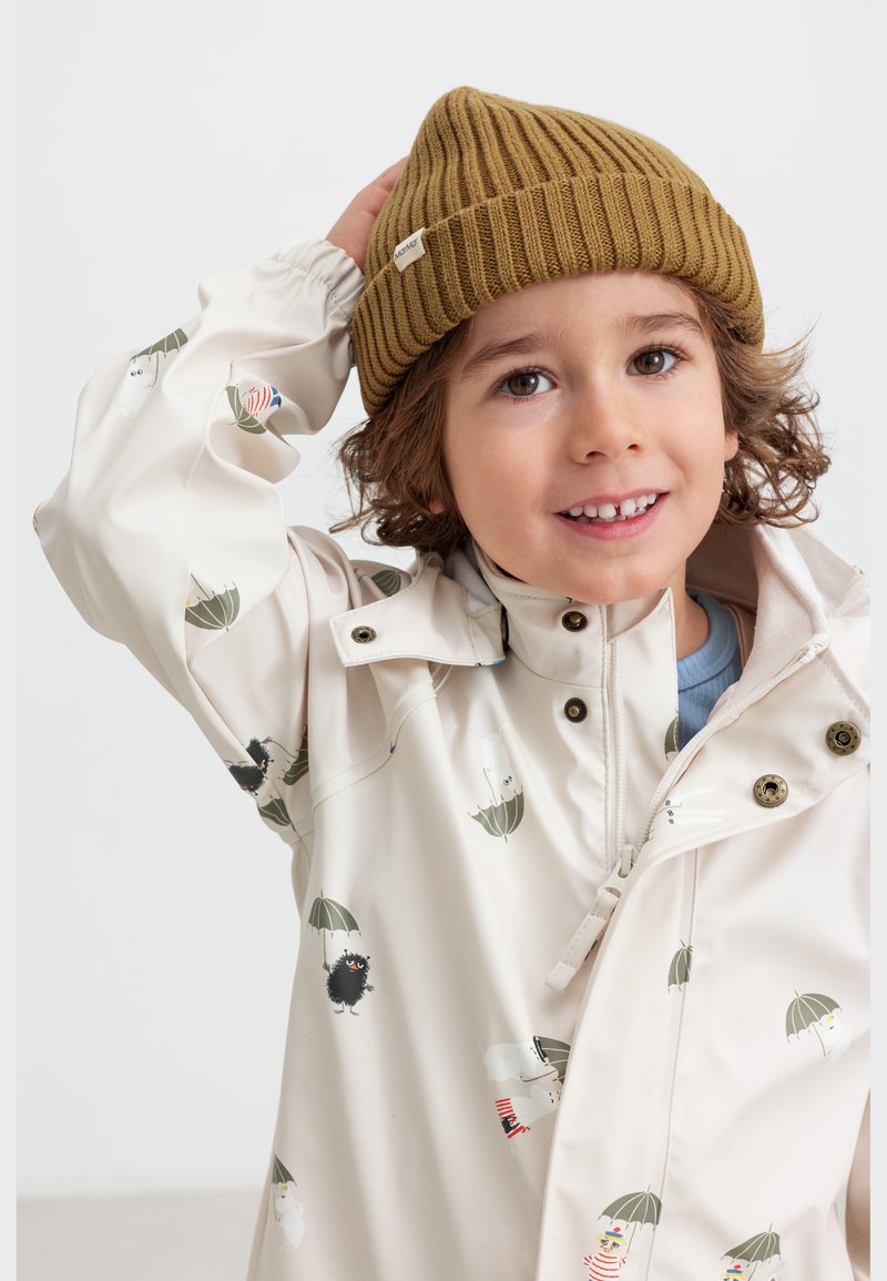 Niño sonriente con cabello rizado, lleva un gorro de punto marrón y un impermeable blanco con dibujos de paraguas y personajes, posando con la mano en el gorro.