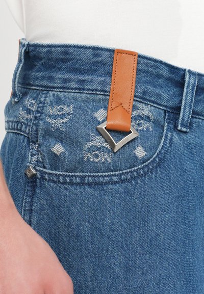 Light blue denim jeans with a brown leather strap and silver hardware, featuring distressed details and embroidered patterns near the pocket.