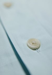 Lichtblauw stoffen shirt met een close-up weergave van een grote, ronde, lichtgekleurde knop, voorzien van een gestikte bovenkant en een gladde textuur.