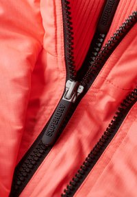 Koral pufferjakke indvendigt med fin tekstureret stof, der har to sorte lynlåse og mærkesynkere.