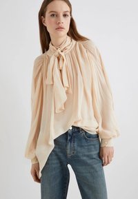 Blusa color pescoce con tessuto morbido, colletto alto e dettaglio a fiocco. Presenta maniche lunghe e spalle leggermente arricciate.