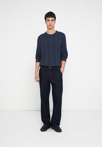 Granatowy sweter w splot warkoczowy z okrągłym dekoltem, zestawiony z ciemnoniebieskimi spodniami o szerokich nogawkach. Model stoi na tle jednolitego białego tła.