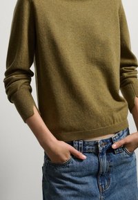 Maglione in lana verde oliva con maniche lunghe, polsini e orlo a coste, abbinato a pantaloni shorts in denim a vita alta. La texture appare morbida e calda.