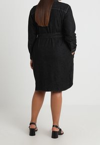 Žena s dlouhými hnědými vlasy, oblečená v černých džínových šatech s páskem a černých otevřených sandálech na blokovém podpatku, stojící s rukama v kapsách.