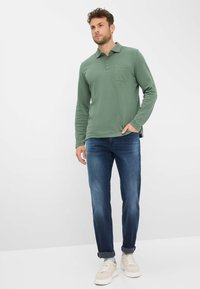 Polo à manches longues, col, de couleur vert olive, avec une poche poitrine, associé à un jean bleu et des baskets beiges.