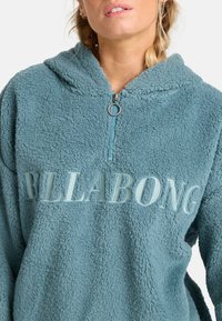 Fleece pullover in teal met een ritssluiting bij de kraag, voorzien van geborduurde "BILLABONG" tekst aan de voorkant en een zachte, gestructureerde oppervlakte.