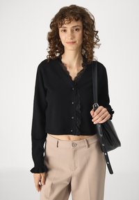 Melnas īsās kardigānas ar mežģīņu apdari ap kaklu un pogu aizdari, valkātas virs bēša augsto jostasvietu biksēm. Melna soma rokā.