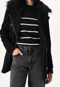 Černý kabát s límcem z umělé kožešiny, přední zip, doplněný černobílým pruhovaným svetrem a šedými džínami z denimu.