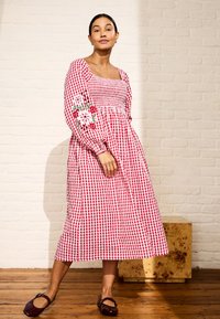 Vrouw in een rood-wit geruite midi-jurk met pofmouwen en geborduurde bloemen, draagt bruine platte schoenen, staand op een houten vloer tegen een witte bakstenen muur.