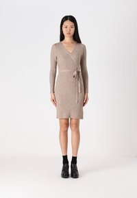 Robe enveloppante en tricot côtelé beige avec des manches longues, un col en V et un détail de ceinture ; silhouette ajustée avec une longueur de jupe au-dessus du genou. Chaussures noires portées.