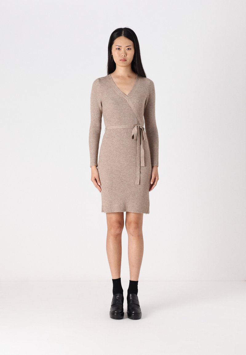 Robe enveloppante en tricot côtelé beige avec des manches longues, un col en V et un détail de ceinture ; silhouette ajustée avec une longueur de jupe au-dessus du genou. Chaussures noires portées.