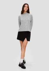 Kobieta stojąca z rękami za plecami, ubrana w szary top z długim rękawem, czarną asymetryczną mini spódnicę, białe skarpetki i czarne loafersy na szarym tle.