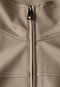 Veste zippée beige avec une texture de tissu lisse, une fermeture éclair foncée contrastante et des panneaux cousus sur le devant pour la structure.