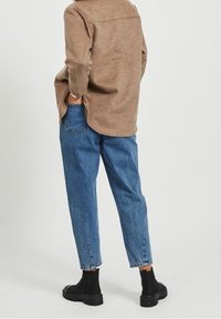 Bruine, gestructureerde shirt met een achterste yoke, gecombineerd met loszittende blauwe jeans en zwarte enkellaarsjes, gezien van achter.