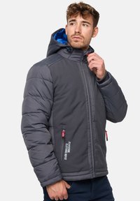 Veste rembourrée grise avec une doublure de capuche bleue, poches zippées, col montant et accents de tirette rouge ; présente une texture matelassée et un design léger.