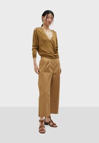 Maglione marrone con scollo a V profondo, maniche lunghe e bottoni sulle spalle, abbinato a pantaloni larghi beige chiaro e sandali marroni con strappi.