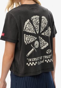 Camiseta negra con un gráfico de pizza en rodajas y un cortapizzas, con el texto "En la corteza confiamos" y "Ve local" en la parte posterior, llevada por una persona con cabello rubio.
