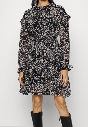 Musta kukkakuvioinen röyhelöinen mekko, jossa pitkät läpikuultavat hihat, vyötärö nyörillä ja polvipituinen helma, puettuna mustien polven korkeiden saappaiden kanssa.