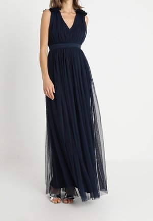 Kobieta w granatowej, długiej do ziemi, plisowanej sukni bez rękawów z dekoltem w kształcie litery V i przezroczystą warstwą, w połączeniu z srebrnymi, odkrytymi na palcach szpilkami.