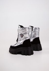 Bottes argentées gonflées, hauteur cheville, avec des accents en suède noir et une semelle noire épaisse et striée. Possède une fermeture éclair et un détail de logo en triangle.
