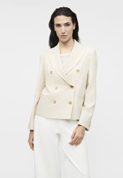 Γυναίκα με σκούρα μαλλιά που φοράει ένα εκρού διπλό ιμάντα σακάκι με καφέ κουμπιά και λευκό παντελόνι, στέκεται μπροστά σε απλό φωτεινό φόντο.