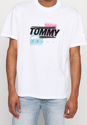 Mężczyzna w białej koszulce z krótkim rękawem z kolorowym logo "TOMMY JEANS" i jasnoniebieskich dżinsach, stojący na jasnym tle.