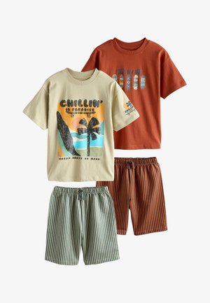 To barefodede drenge står indendørs iført T-shirts med grafik og stribede shorts i et lyst rum med sofa og vægkunst bag dem.