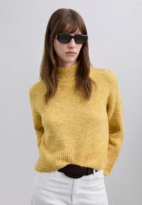 Pull à col roulé côtelé jaune avec une finition texturée, associé à un jean blanc et une ceinture foncée. Le modèle porte des lunettes de soleil marron.