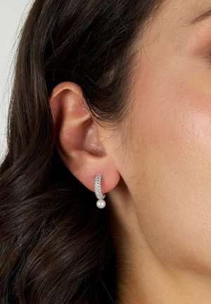 Orecchio di donna con capelli scuri che indossa un piccolo orecchino a cerchio d'argento decorato con pietre trasparenti e una perla pendente.