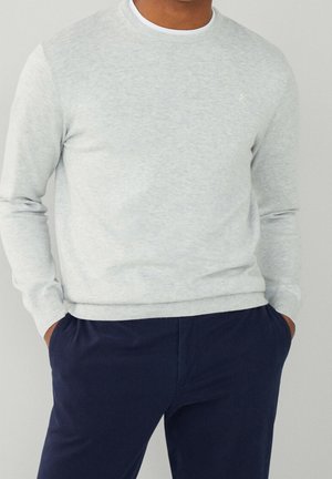 Homme portant un pull gris clair à col rond et un pantalon bleu marine foncé, les mains dans les poches, sur fond uni.